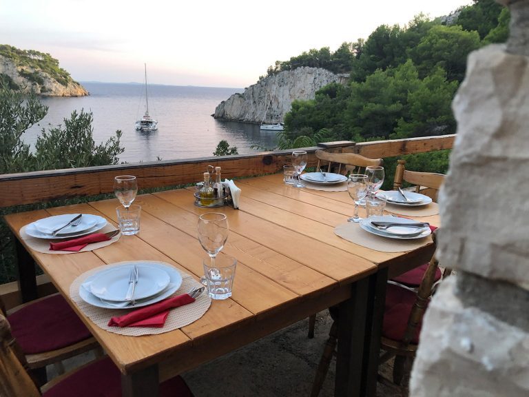 Gedeckter Holztisch auf Terrasse mit Blick auf eine Bucht in Kroatien, in der Segelboote vor Anker liegen.