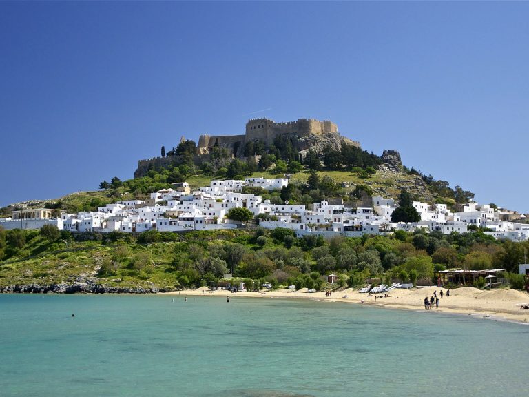 Weiße Häuser von Lindos und die Akropolis auf Rhodos erheben sich über einer Bucht mit türkisfarbenem Wasser und Sandstrand.