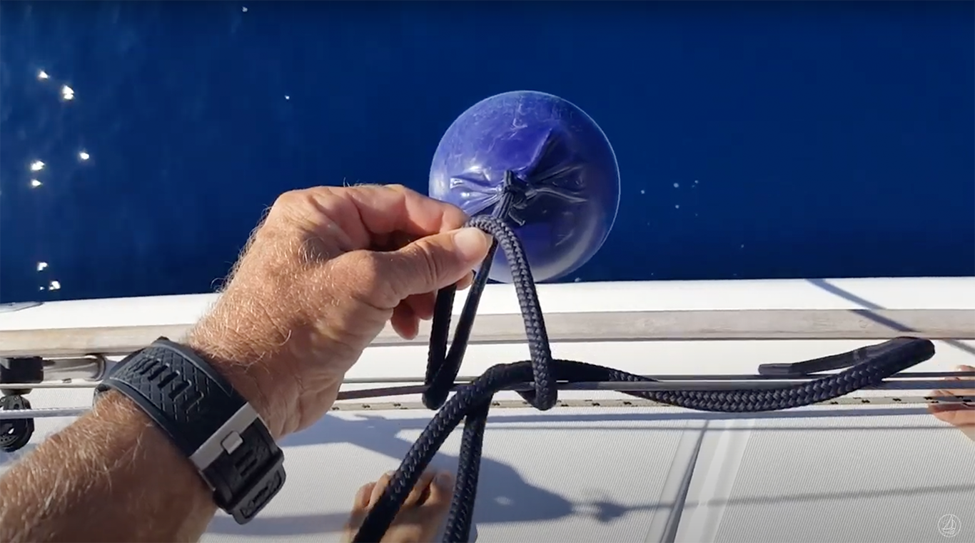 Hand knüpft einen Knoten in eine Leine an einer blauen Kugelboje an Bord eines Segelboots.