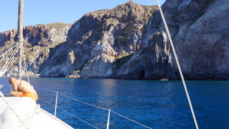 Segelboot ankert vor steilen Felsklippen an der Küste zwischen Mallorca und Sardinien, eine Person liegt am Bug.