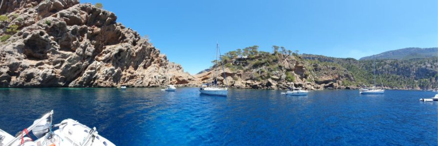 Segelboote ankern in einer felsigen Bucht vor Mallorca mit türkisblauem Wasser und bewaldeten Klippen.