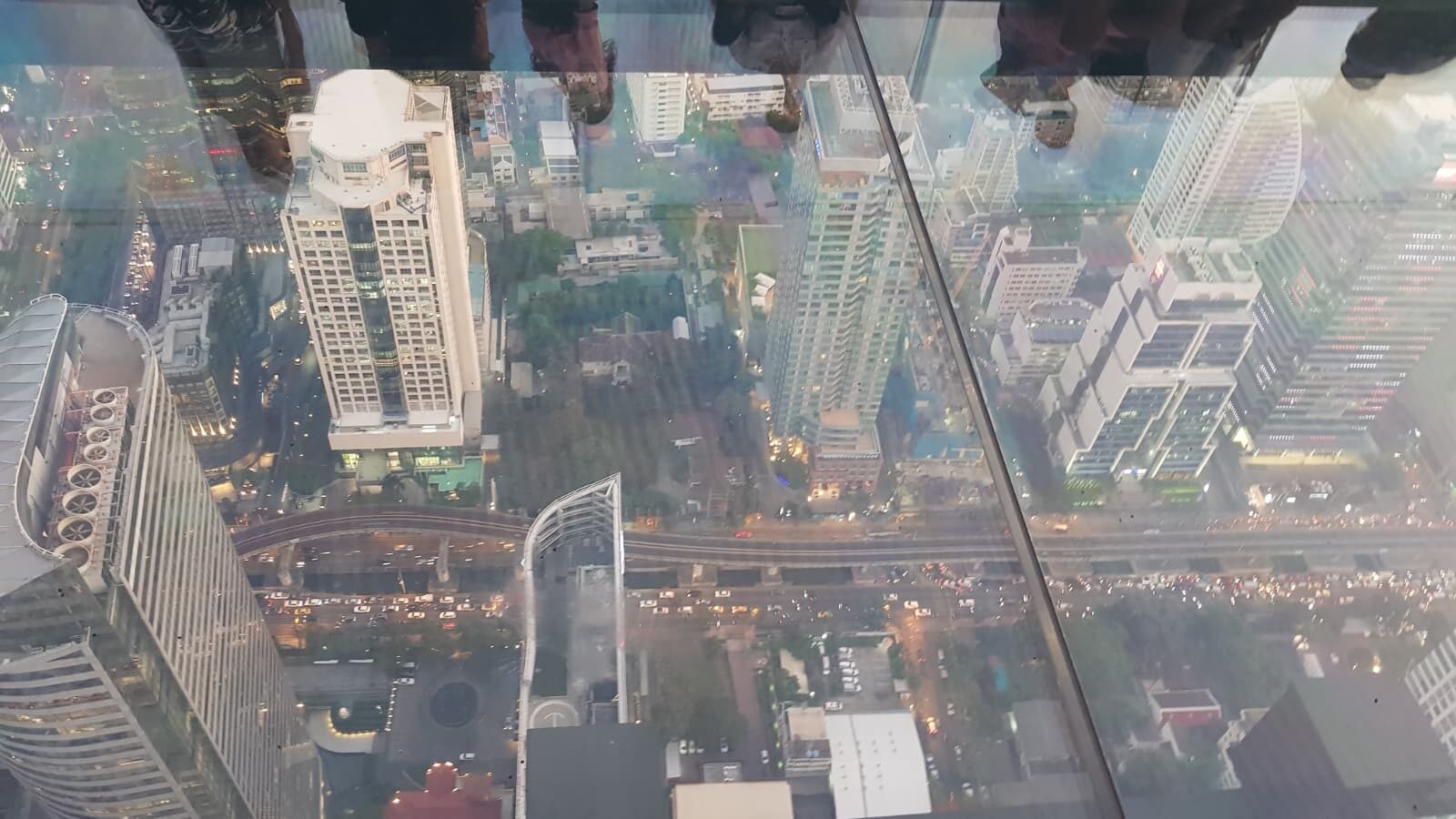 Blick durch Glasboden auf Wolkenkratzer, Straßenverkehr und Grünflächen von Bangkok von oben.