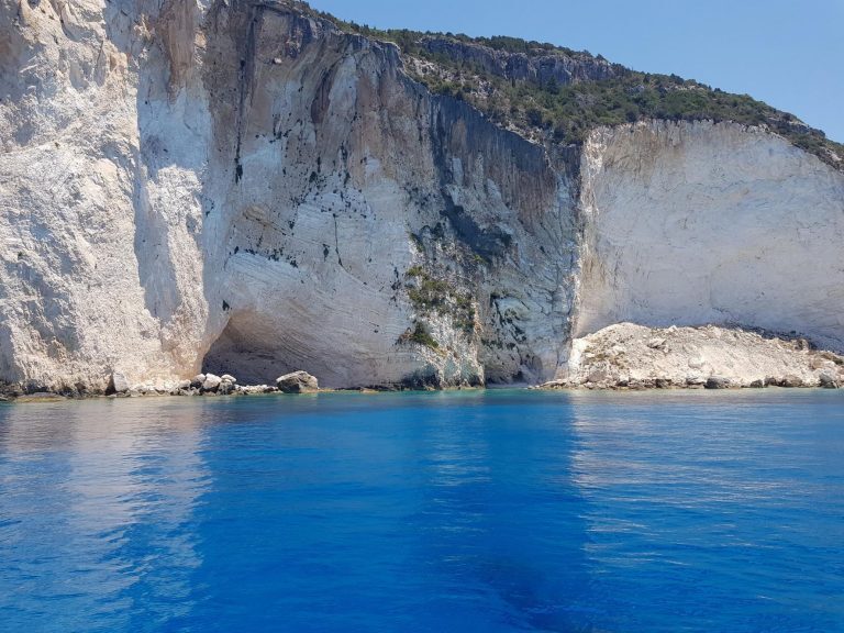 Steile Kalksteinklippen fallen an der griechischen Küste ins tiefblaue Meer, oben bewachsen mit grünen Büschen.