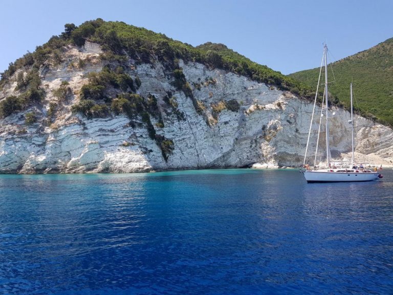 Segelboot vor einer bewaldeten Felsküste im türkisblauen Wasser des Ionischen Meeres.