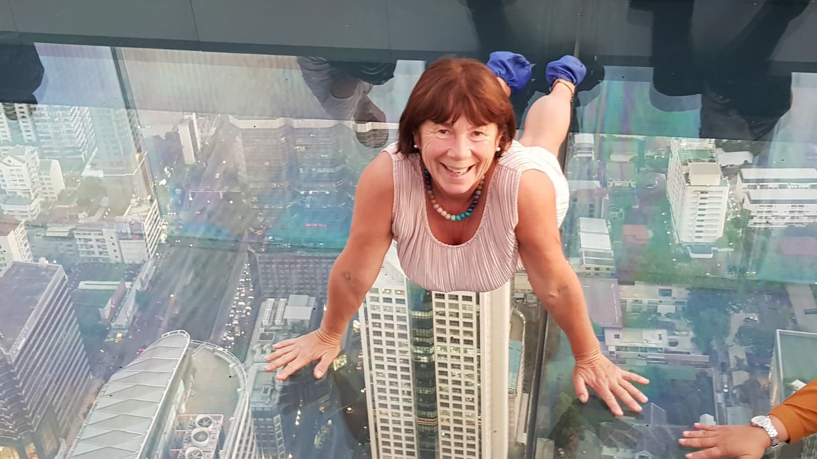 Frau posiert lächelnd auf Glasboden-Skywalk hoch über der Skyline von Bangkok.