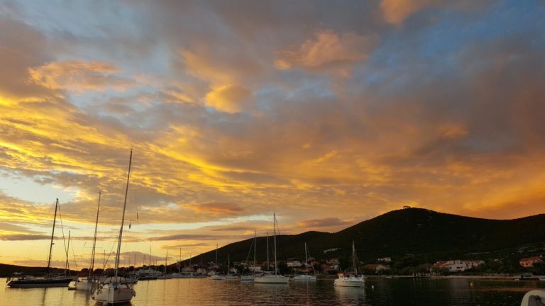 Segelboote liegen in einer Bucht an der kroatischen Küste vor einem Hügel unter orange gefärbtem Wolkenhimmel.