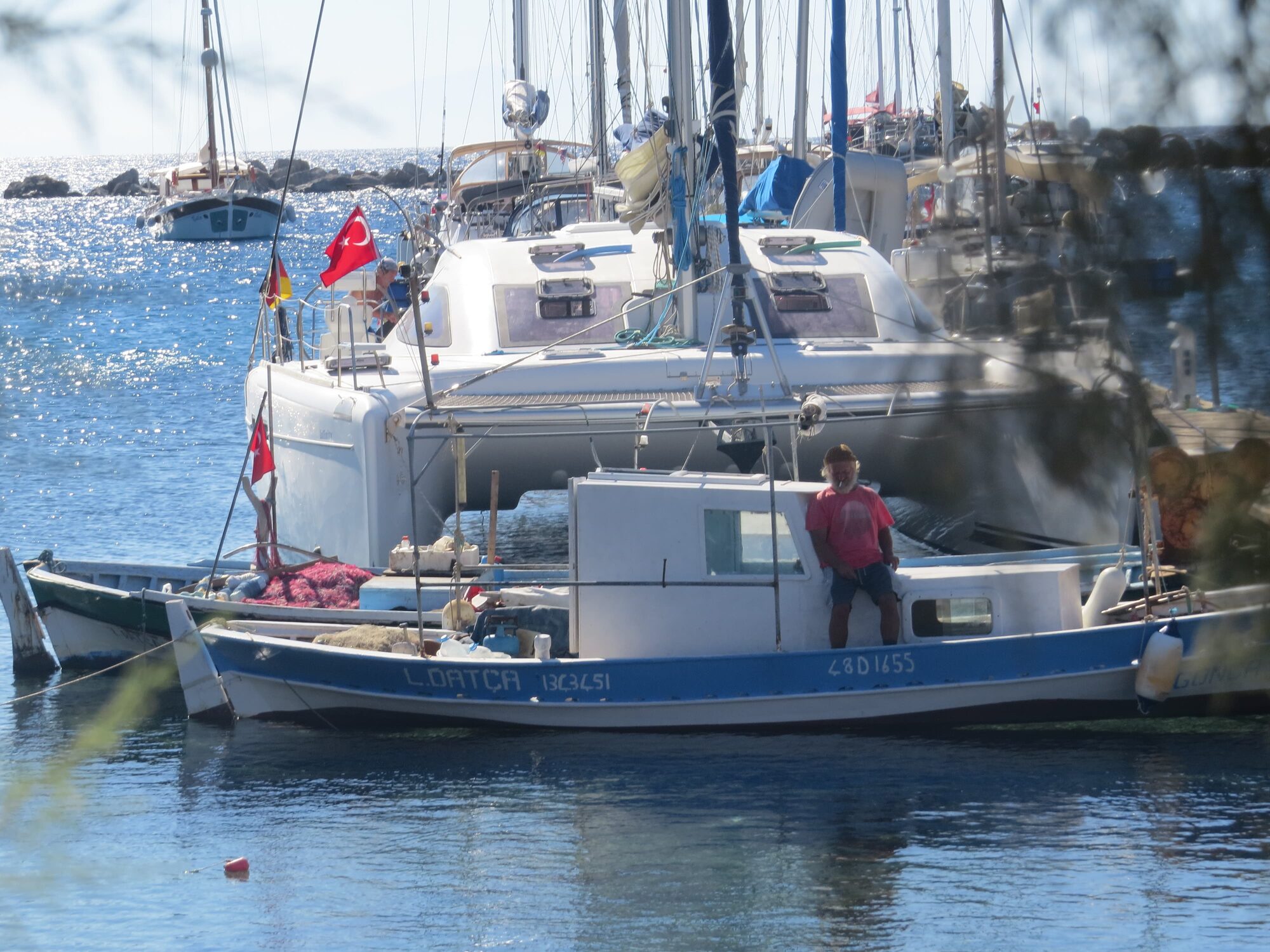 Fischerboot „L.Datça“ mit Mann im Hafen, dahinter Segelyachten mit türkischen Flaggen, Ägäis, Türkei.