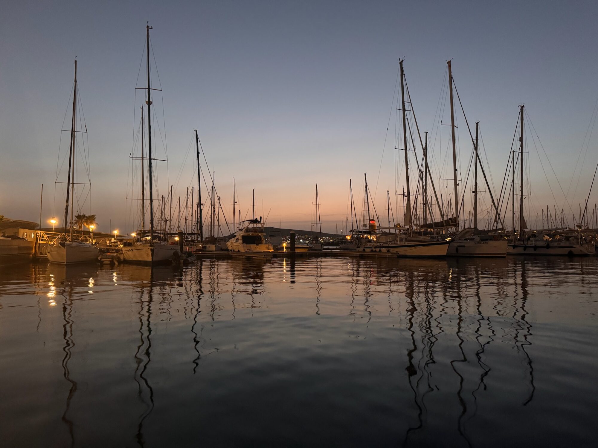Segelboote mit hohen Masten liegen in der Marina Rubicón auf Lanzarote, ihre Silhouetten spiegeln sich im Wasser bei Dämmerung.