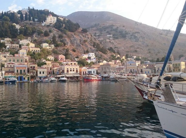 Bunte Häuser am Hafen von Symi im Dodekanes, davor Segelboote im Wasser vor bergiger Kulisse.