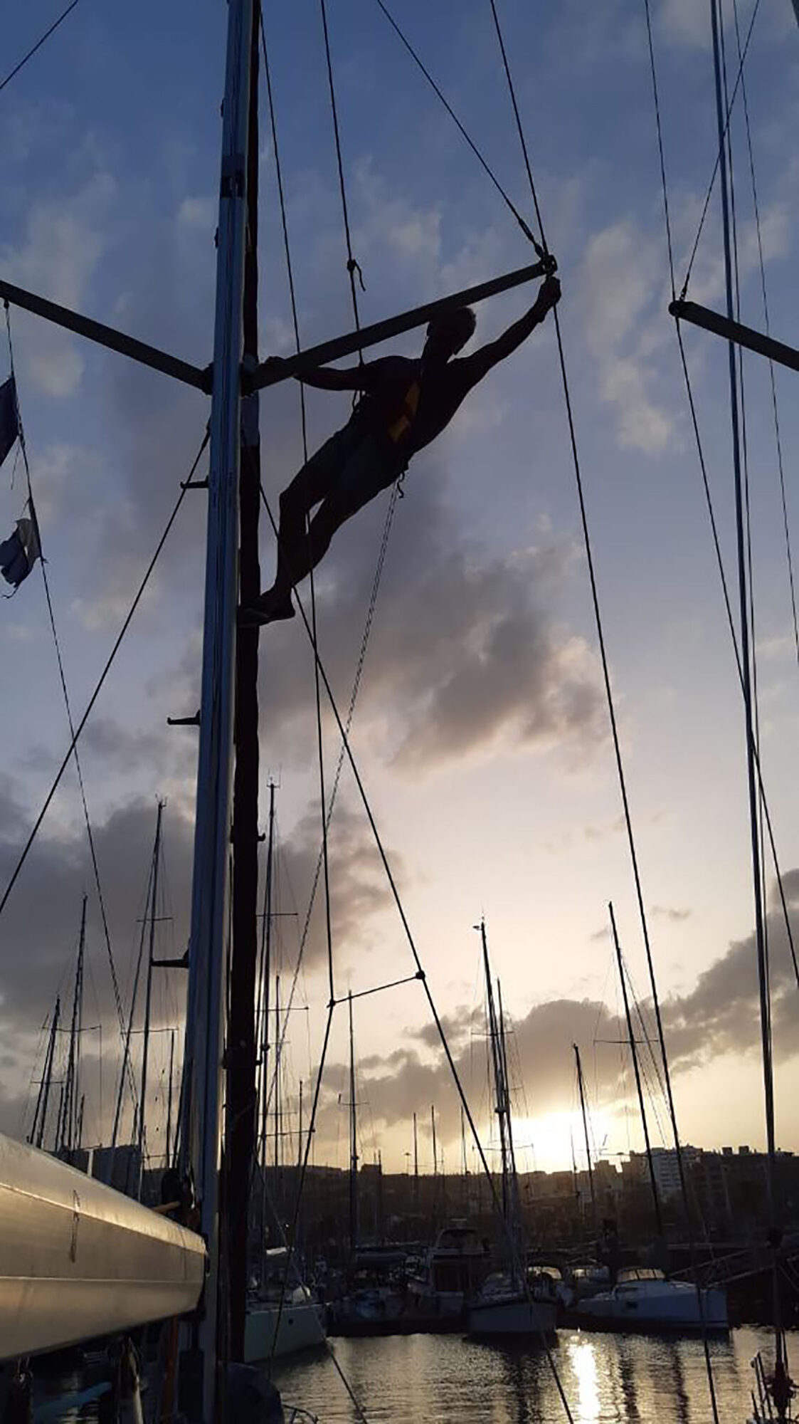 Eine Person klettert im Mittelmeerhafen bei Sonnenuntergang am Mast eines Segelboots zwischen den Masten anderer Boote.