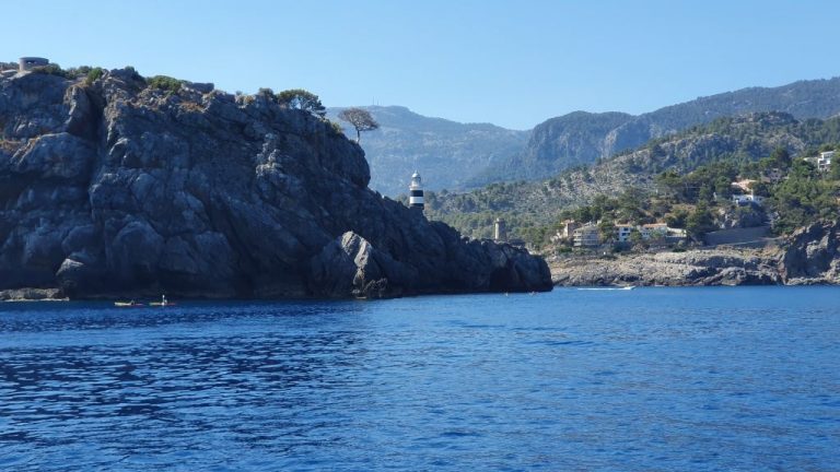Felsige Küste Mallorcas mit Leuchtturm an der Hafeneinfahrt, Bergen im Hintergrund und Kajaks auf dem Wasser.