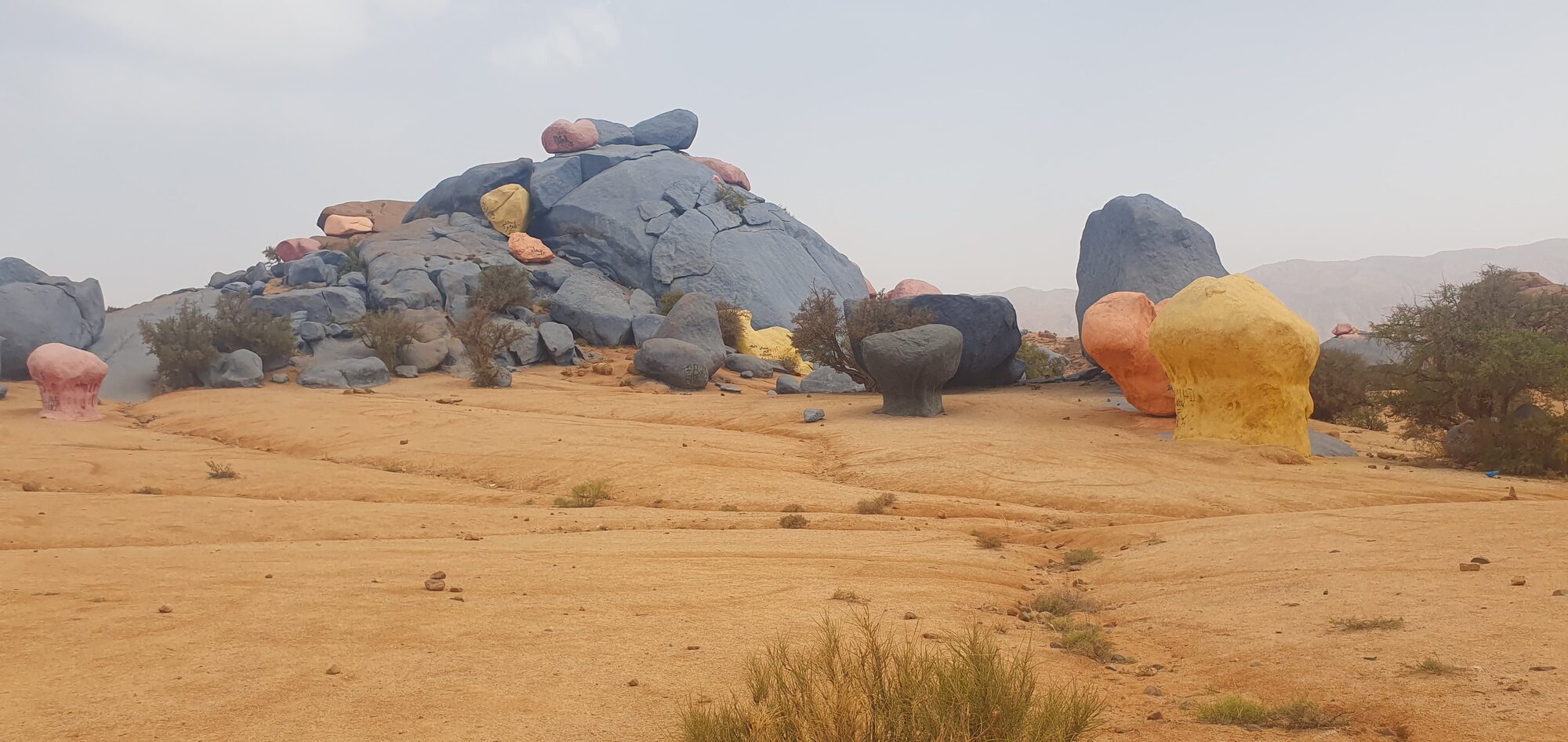 Bunt bemalte Felsbrocken und ein Steinhaufen in sandiger Wüstenlandschaft mit vereinzelten Büschen, Marokko.