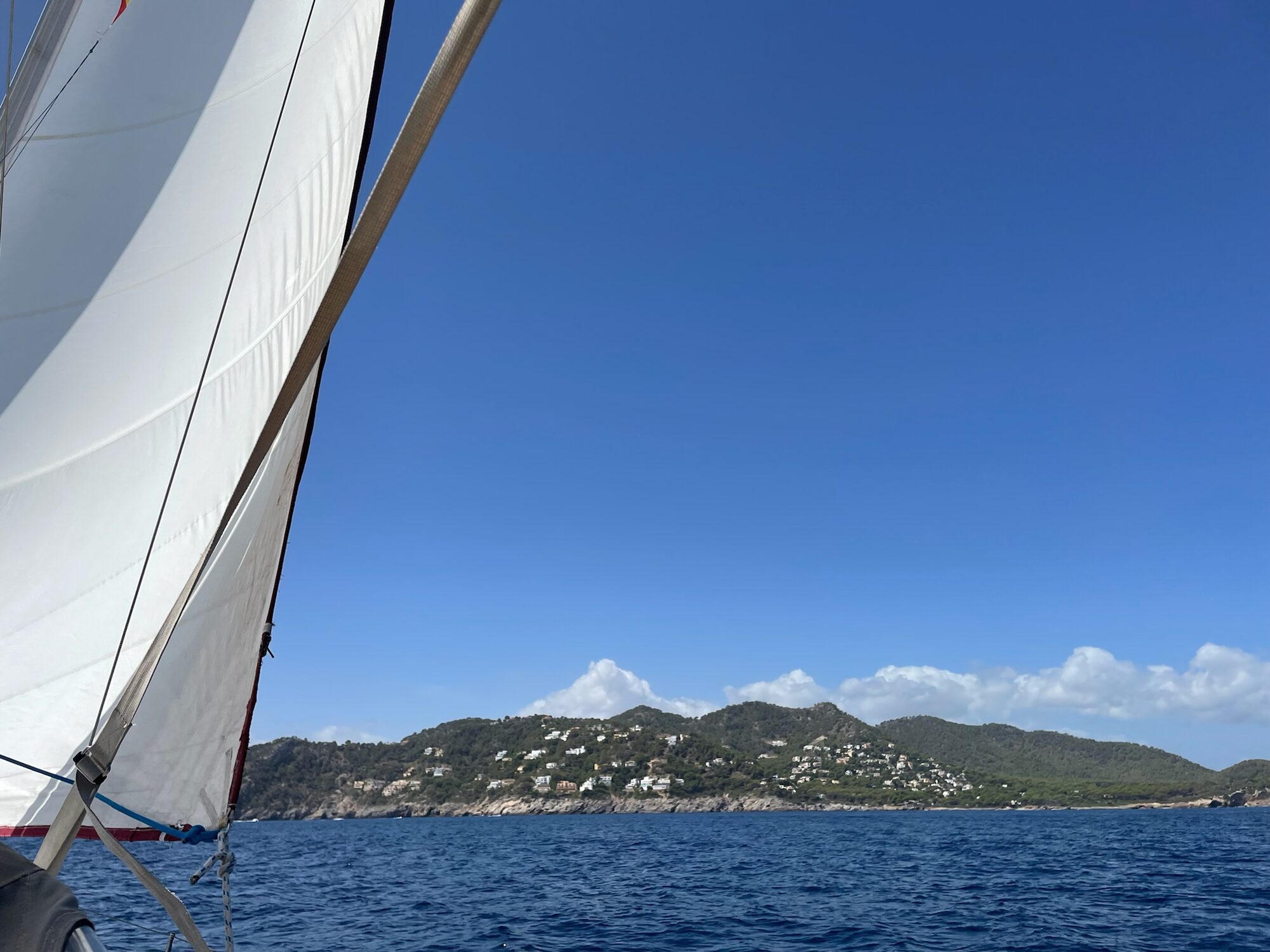Segelboot vor der bewaldeten Küste Mallorcas mit verstreuten Villen an einem Hang über dem Meer.