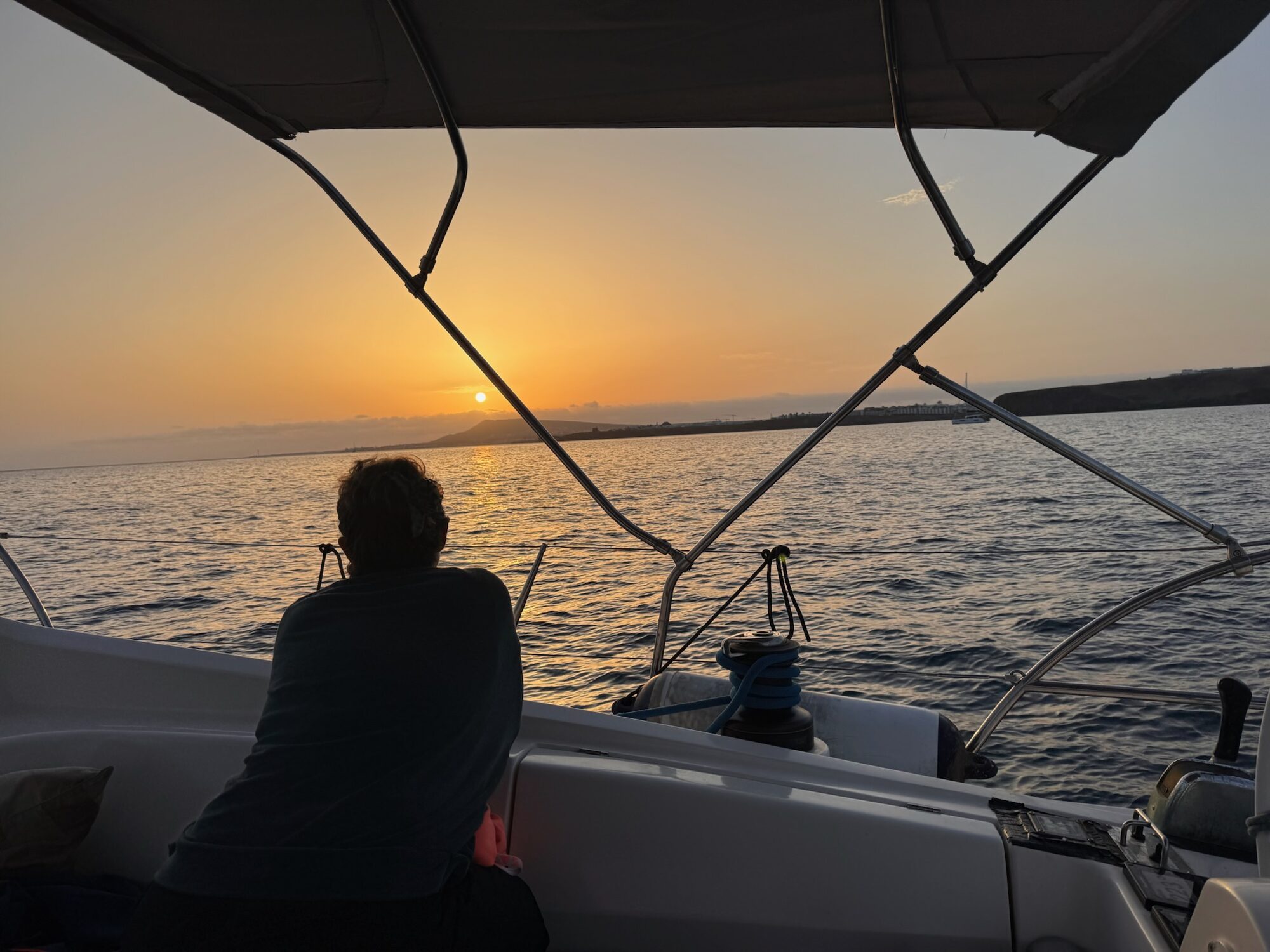 Person sitzt im Cockpit eines Segelboots vor Lanzarote und blickt bei Sonnenuntergang auf die Küste.
