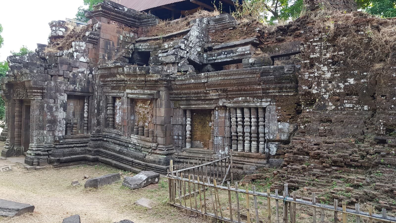 Verwitterte Sandsteinruine eines Khmer-Tempels mit verzierten Fenstern und Säulen, überwuchert von Moos, hinter einem Bambuszaun.