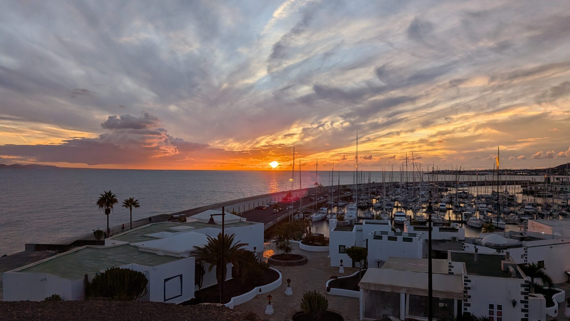 Sonnenuntergang über der Marina Rubicón auf Lanzarote, mit weißen Häusern, Palmen und zahlreichen Segelbooten am Atlantik.