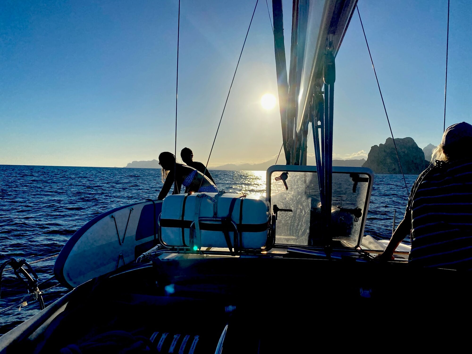 Menschen auf einem Segelboot mit Stand-up-Paddle-Board bei Sonnenuntergang, im Hintergrund Felsküste im Meer.