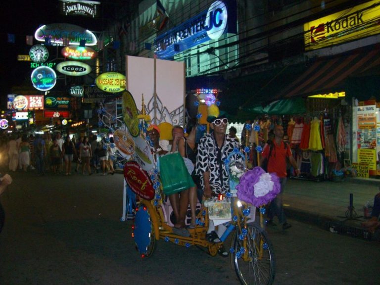 Bunt geschmücktes Fahrrad-Taxi fährt nachts durch eine belebte, mit Leuchtreklamen gesäumte Straße in Thailand.
