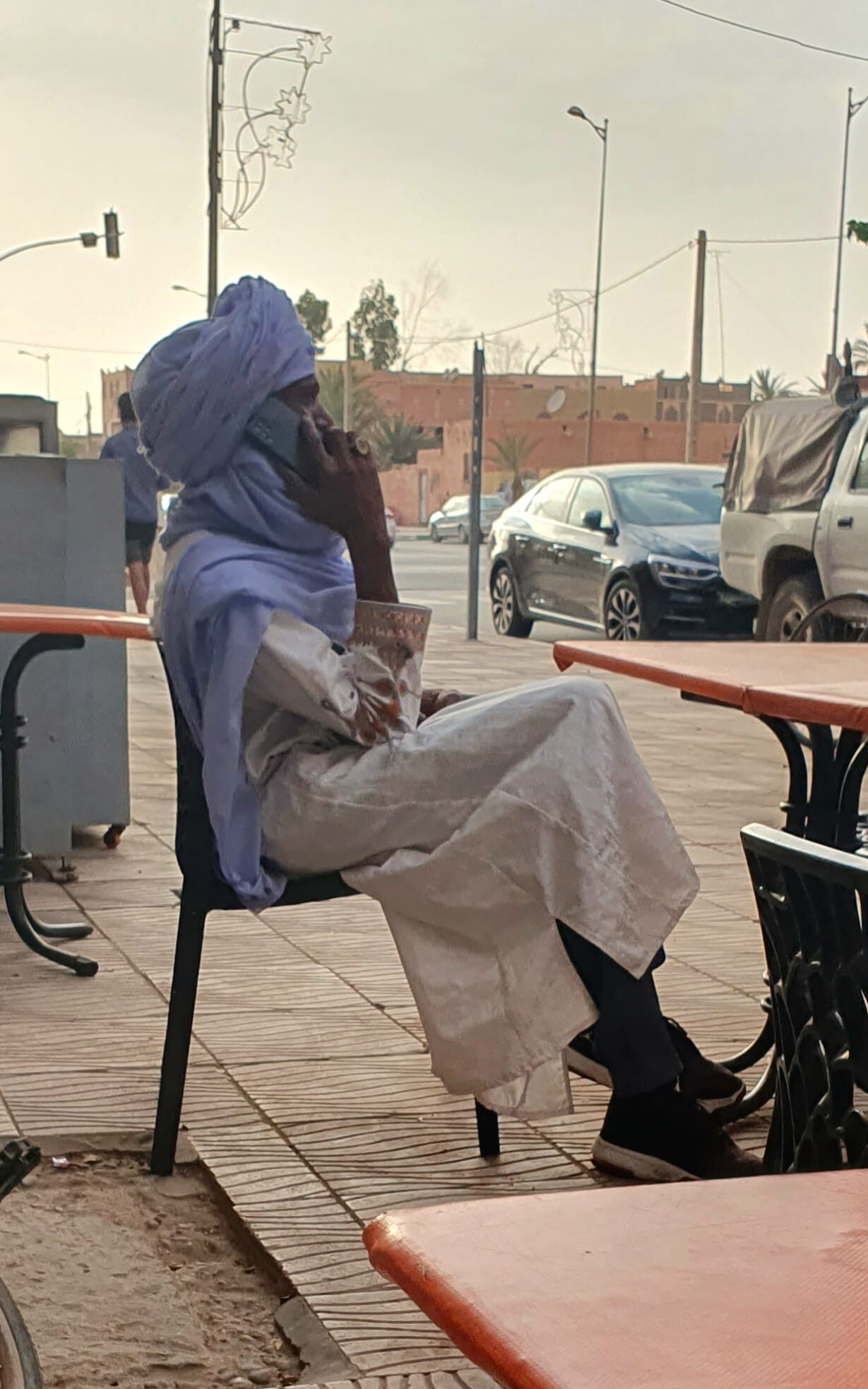 Mann in traditioneller blauer Tuareg-Tracht sitzt telefonierend auf einem Plastikstuhl an einem Straßencafé in Marokko.