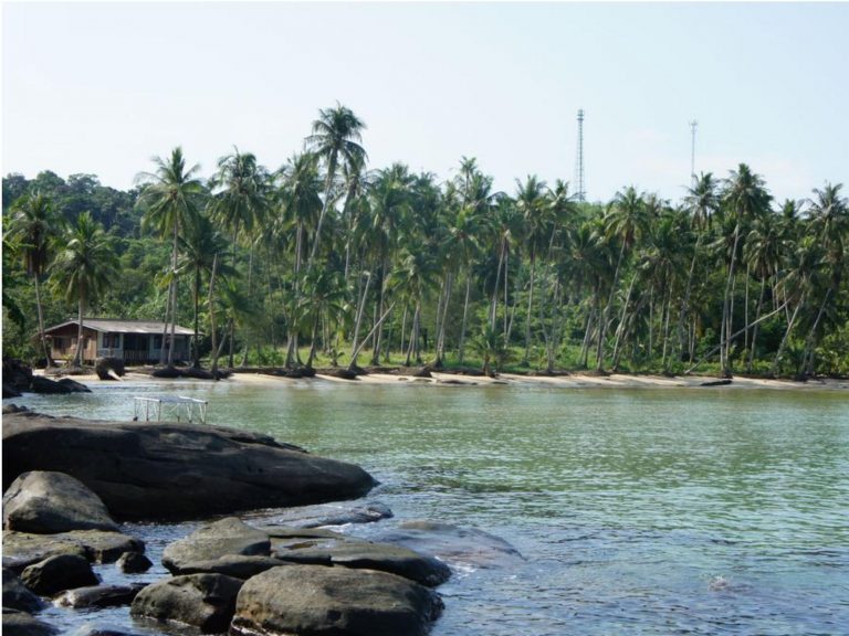 Palmenbestandene Küste mit Sandstrand, Felsen im Vordergrund und einer Hütte am Ufer in der Phang-Nga-Bucht, Thailand.
