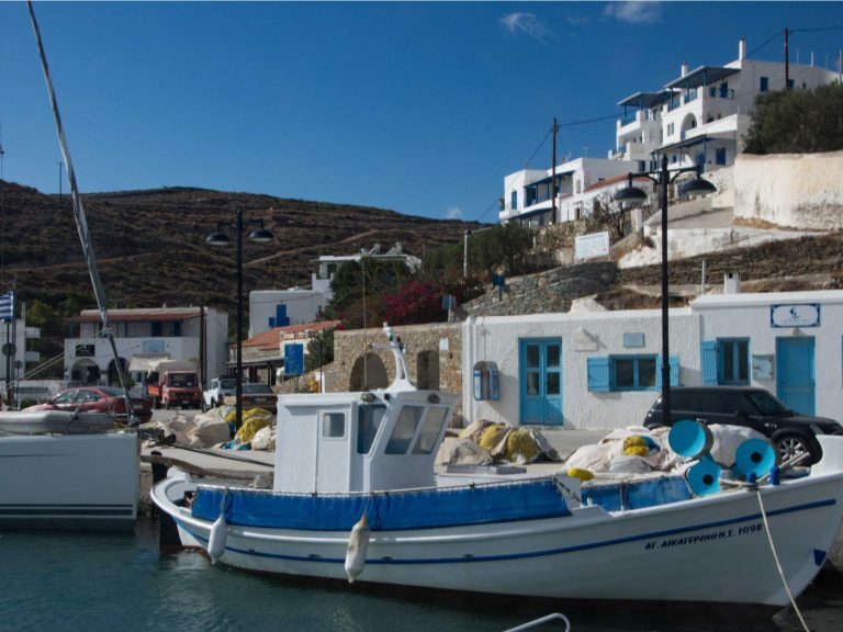 Fischerboot liegt im Hafen eines kykladischen Dorfes mit weißen Häusern am Hang, unter blauem Himmel.