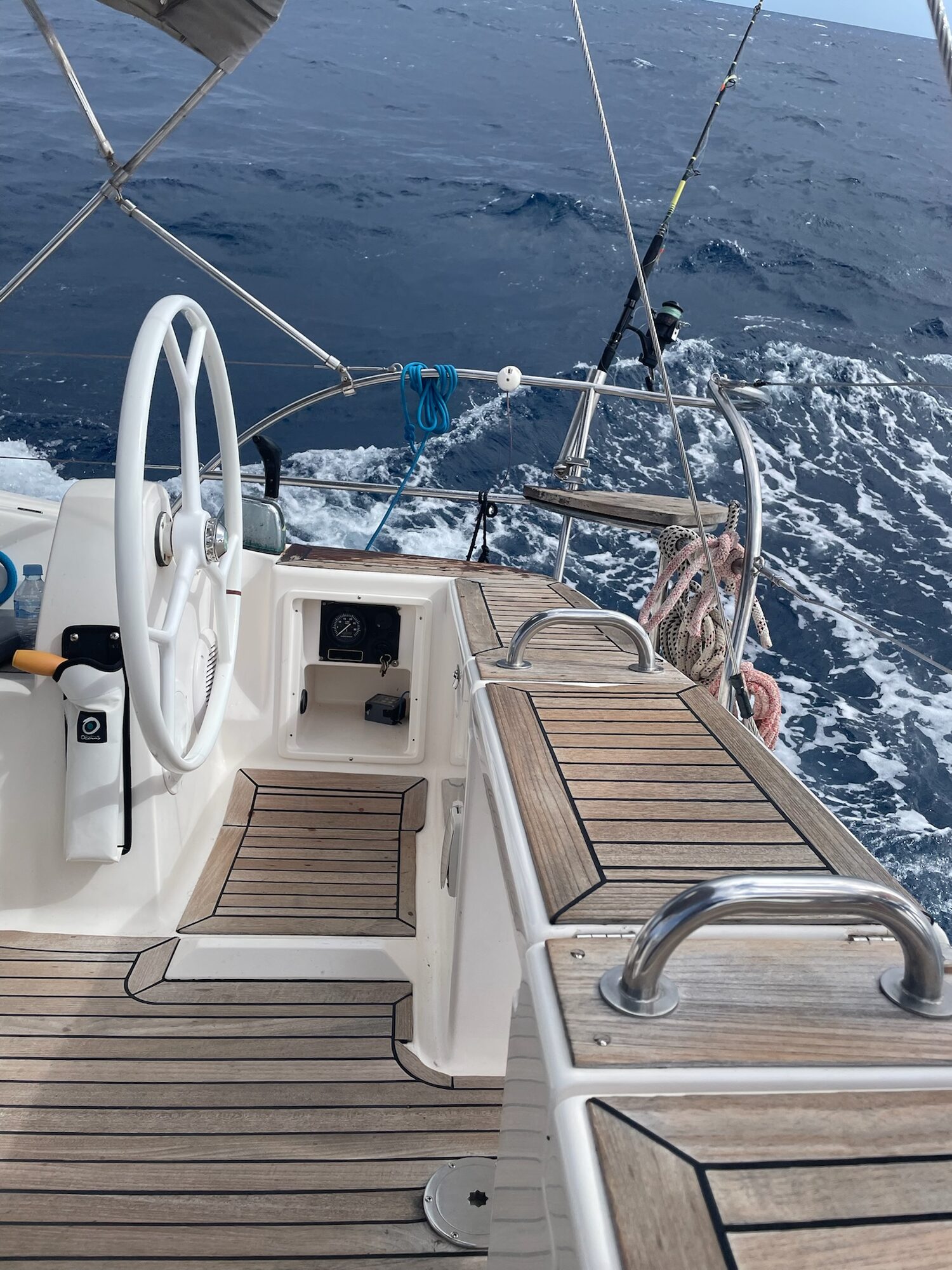Steuerrad und Holzdeck eines Segelboots auf hoher See mit Wellen und Gischt rundherum.