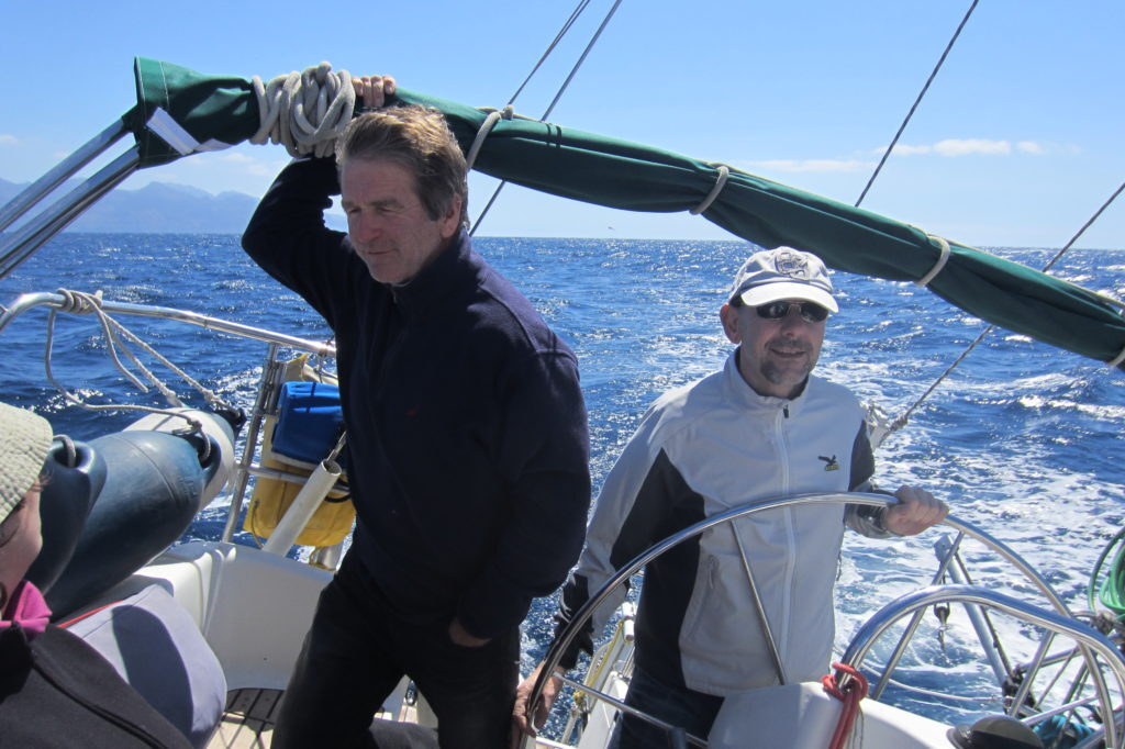 Zwei Männer stehen im Cockpit eines Segelboots, einer hält das Steuerrad, im Hintergrund Meer und Küstenberge.