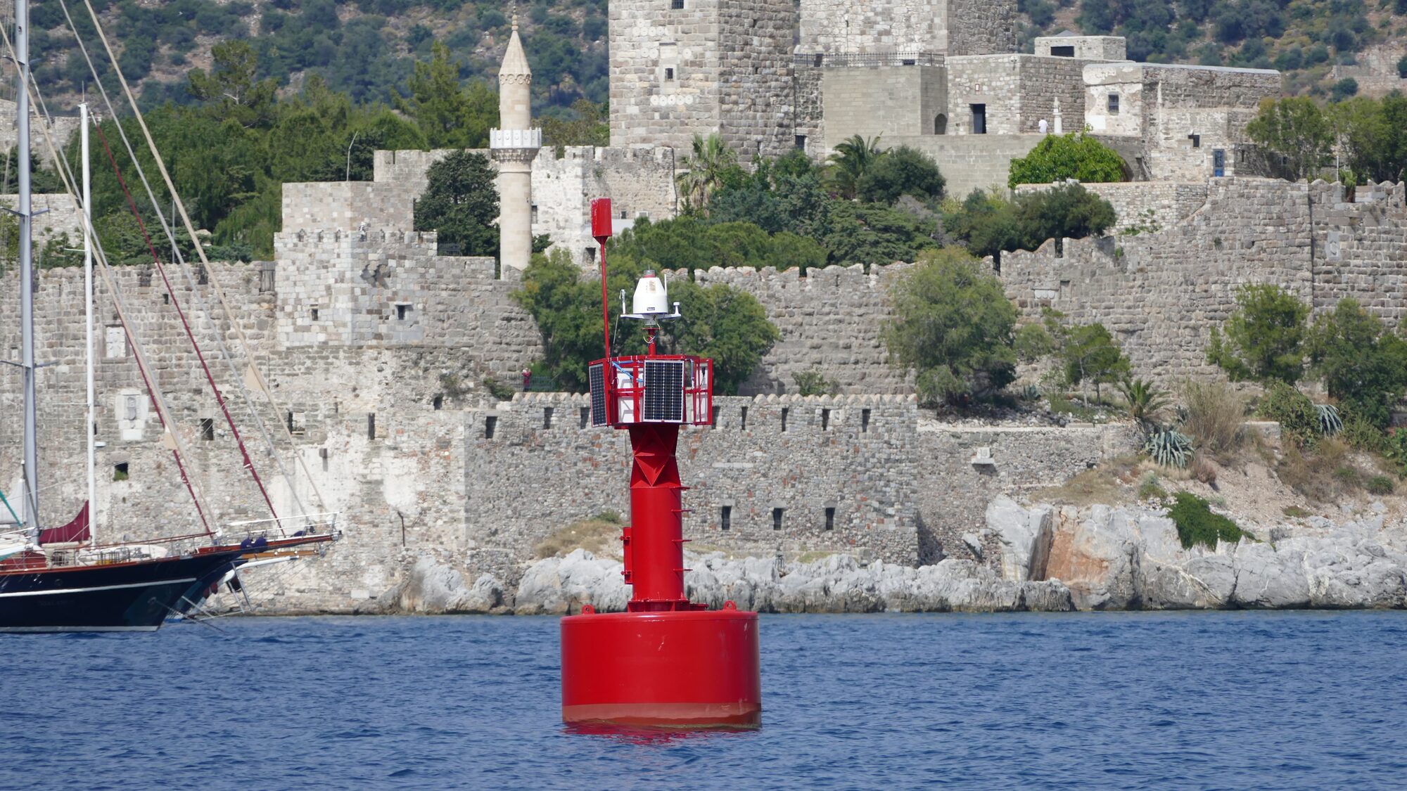 Rote Fahrwassertonne mit Solarpaneelen vor der Festung von Bodrum an der Ägäis, im Vordergrund ein Segelschiff.