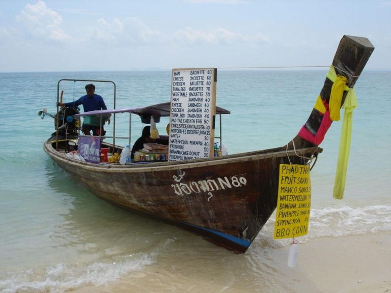 Longtail-Boot mit Snackbar und Speisetafeln liegt am Sandstrand von Krabi, im Hintergrund türkisfarbenes Meer.