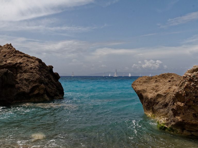 Felsbuchten säumen das türkisfarbene Meer vor Rhodos, im Hintergrund kreuzen mehrere Segelboote auf der Ägäis.