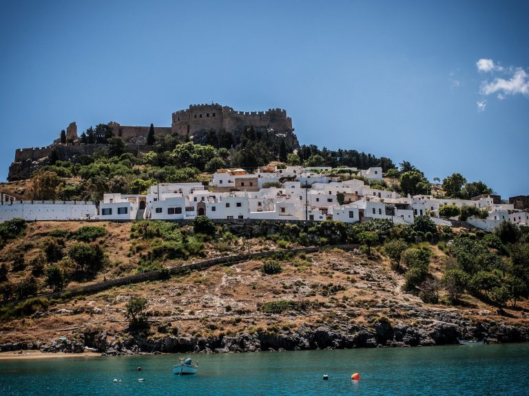 Weiße Häuser von Lindos auf Rhodos unterhalb der antiken Akropolis, davor türkisfarbenes Meer mit einem Boot.