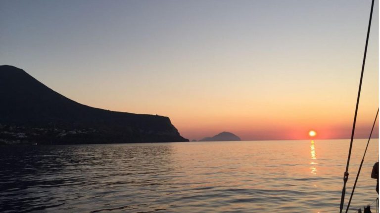 Sonnenuntergang über einer Küstenlandschaft mit vorgelagerten Inseln, gesehen von einem Segelboot aus.
